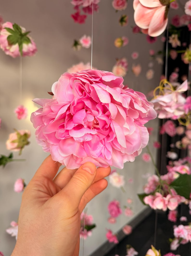 May include: A large, vibrant pink peony flower with layered petals is held in a hand. The background features a soft focus of other pink flowers, creating a floral theme.