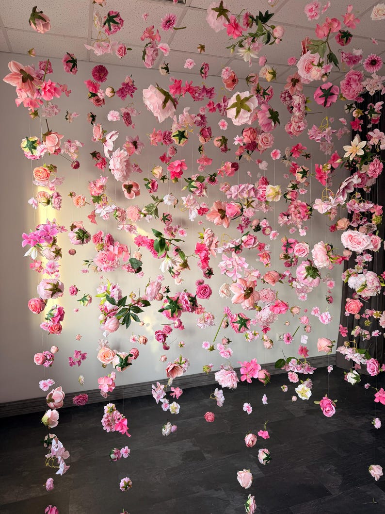 A display of pink and white flowers suspended from the ceiling. The flowers are of various types and sizes, creating a whimsical and romantic atmosphere. The flowers are arranged on strings, creating a curtain-like effect. Hanging flowers romantic decor, silk floating flower garland, floral party backdrop