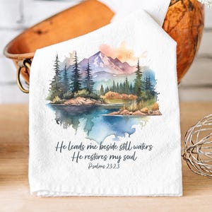 May include: White kitchen towel with a watercolor painting of a mountain lake scene. The text "He leads me beside still waters He restores my soul Psalms 23:23" is printed below the image.