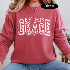 May include: Crimson-colored long-sleeve sweatshirt with the text "BY HIS GRACE Romans 3:24" in white block letters. The sweatshirt has a crew neck and a relaxed fit. The image shows a close-up of the front of the sweatshirt.