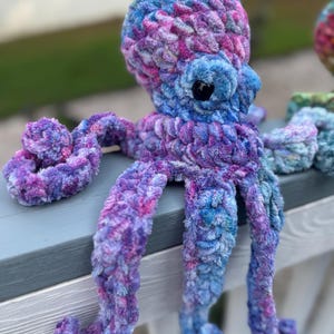 May include: A handmade plush octopus in shades of blue, purple, and pink. The soft, textured toy has large, round eyes and eight long, curled tentacles. The octopus is sitting on a gray surface.