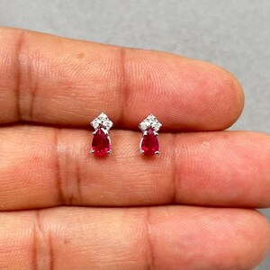 May include: A pair of stud earrings with a teardrop-shaped, deep red gemstone, topped with a cluster of small, clear stones. The earrings are set in silver-coloured metal, presenting a classic and elegant design, suitable for various occasions.