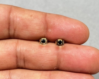 Könnte beinhalten: Ein Paar kleiner, goldfarbener Ohrstecker. Jeder Ohrring hat einen quadratischen, schwarzen Edelstein, der in einen quadratischen Goldrahmen gefasst ist. Die Ohrringe werden vor einem neutralen Hintergrund präsentiert.
