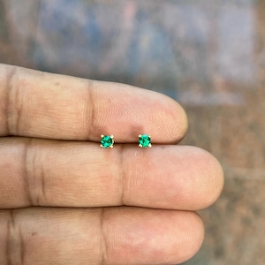 May include: A pair of small, gold-colored stud earrings, each featuring a vibrant green gemstone. The earrings are set in a simple, square-shaped setting, and are displayed against a blurred background.