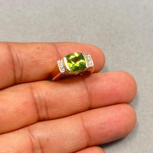 May include: A gold ring featuring a large, square-cut, light green gemstone. The ring has small, rectangular accents with clear stones on each side of the main stone. The ring is held in a hand against a gray background.