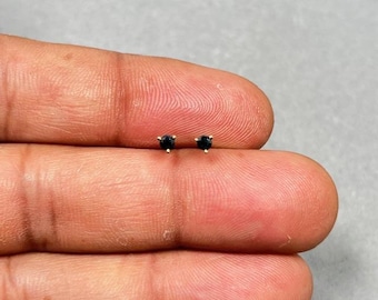 Könnte beinhalten: Ein Paar kleiner Ohrstecker mit einem dunkelblauen Edelstein. Die Ohrringe haben einen goldfarbenen Stift und eine Fassung. Die Edelsteine sind rund und haben einen Durchmesser von etwa 0,25 cm. Die Ohrringe werden auf hellem Hintergrund präsentiert.