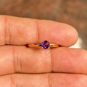 May include: A gold-colored ring with a round, deep purple gemstone. The ring's band is thin and polished, and the gemstone is set in a four-prong setting. The ring is held between fingers, showcasing its design.