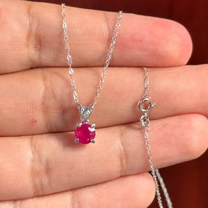 Può includere: Una delicata collana d'argento con un vivace pendente di rubino rotondo. Il rubino è incastonato in una montatura dettagliata e ornata. La catena ha un design fine e ritorto con una chiusura sicura. La collana è mostrata su uno sfondo morbido e neutro.