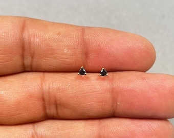 Könnte beinhalten: Ein Paar kleiner Ohrstecker mit schwarzen, dreieckigen Edelsteinen, die in goldfarbenem Metall gefasst sind. Die Ohrringe werden auf hellem Hintergrund präsentiert.