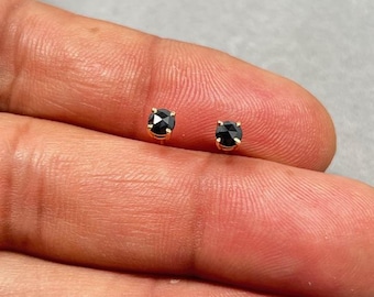 Könnte beinhalten: Ein Paar kleiner, runder Ohrstecker mit schwarzen Edelsteinen und goldfarbenen Fassungen. Die Ohrringe werden auf einer hellgrauen Oberfläche präsentiert. Die Edelsteine haben mehrere Facetten, die das Licht reflektieren.