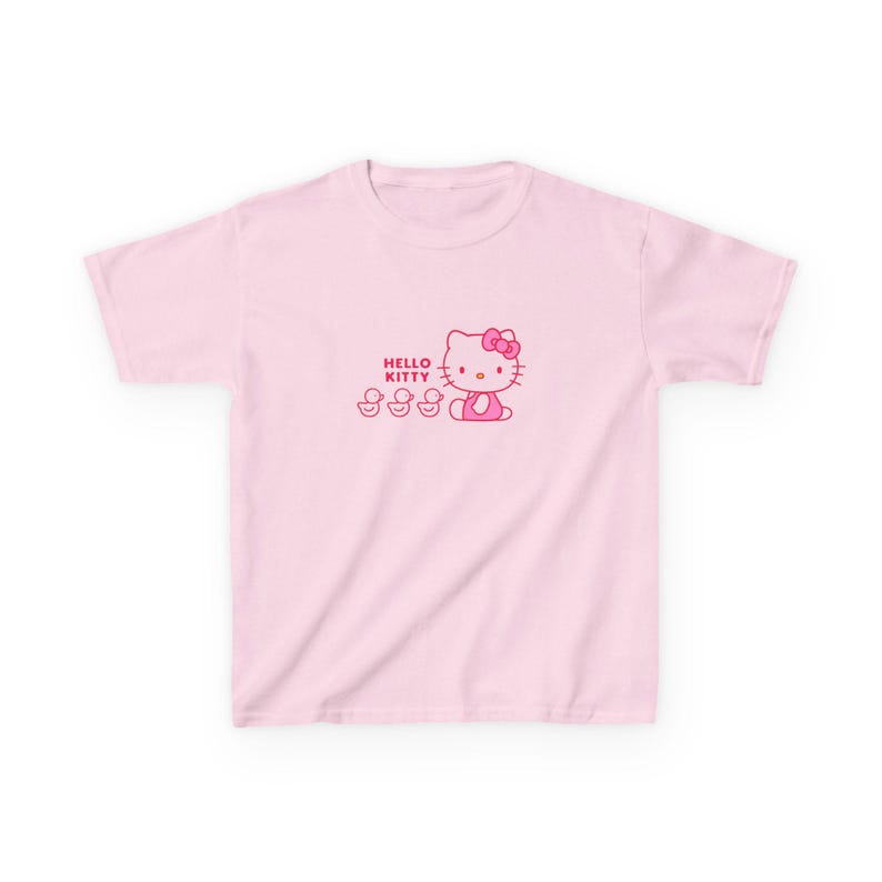 Puede incluir: Camiseta rosa claro con un dise&ntilde;o de Hello Kitty. El dise&ntilde;o incluye el texto "HELLO KITTY", Hello Kitty y tres patitos, todos en rosa. La camiseta tiene cuello redondo y mangas cortas.