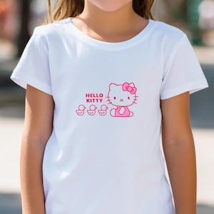 Puede incluir: Camiseta blanca con un gr&aacute;fico rosa de Hello Kitty con un lazo y tres patitos. Las palabras "Hello Kitty" est&aacute;n impresas encima del personaje. La camiseta tiene mangas cortas y cuello redondo.