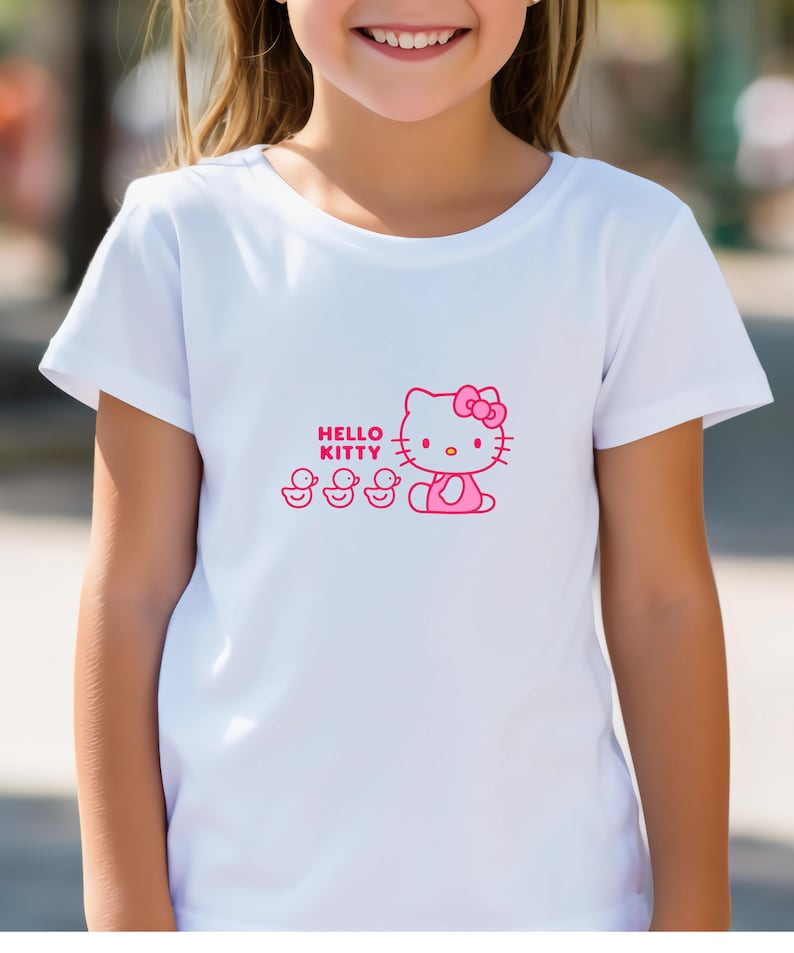 Puede incluir: Camiseta blanca con un gr&aacute;fico rosa de Hello Kitty con un lazo y tres patitos. Las palabras "Hello Kitty" est&aacute;n impresas encima del personaje. La camiseta tiene mangas cortas y cuello redondo.