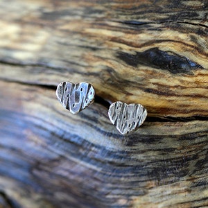 May include: Two silver heart-shaped earrings with a textured surface. The earrings are on a wooden background.