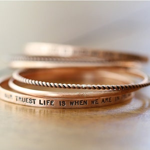 May include: Set of four copper bangles with a rope design. The bottom bangle has the inscription "Our truest life is when we are in the"