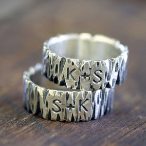 May include: Two silver rings with a textured, bark-like design. Each ring has initials engraved, one with "K+S" and the other with "S+K". The rings are stacked on top of each other, showcasing the unique, handcrafted style.