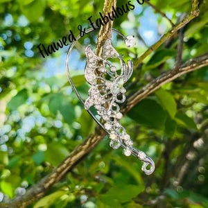 May include: A silver-toned ear cuff with a butterfly design, adorned with clear crystals. The cuff is attached to a curved wire that wraps around the ear. The text "Flawed & Fabulous Co" is visible in the image.