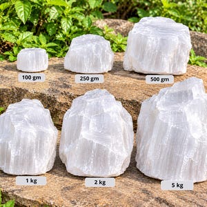 Op de afbeelding: Zes ruwe witte seleniet kristalbrokken gerangschikt in twee rijen. De stenen zijn gelabeld met gewichten van 100 gm, 250 gm, 500 gm, 1 kg, 2 kg en 5 kg. Elk kristal heeft een doorschijnende, vezelige en grillige kristallijne structuur.