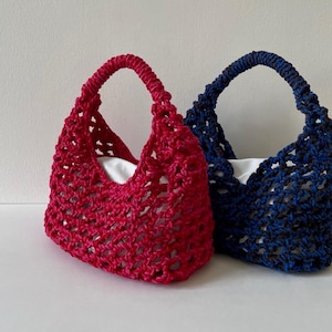 May include: Two handmade crochet bags, one in red and one in blue, with white linings. The bags feature an open weave design and sturdy handles. The text "mühely GIN BAG" is displayed at the top. The size is 20 x 35 x 35 cm.