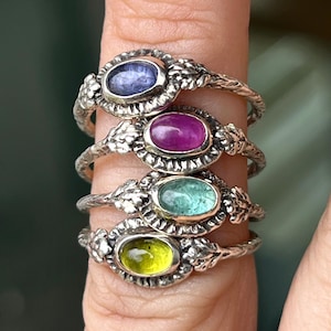 Puede incluir: Cuatro anillos de plata con piedras preciosas de diferentes colores. Los anillos están apilados en un dedo y presentan un delicado diseño floral.