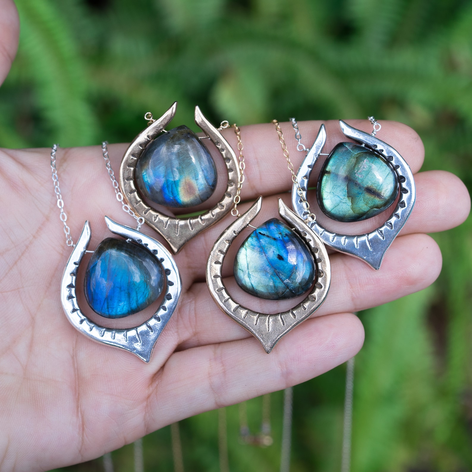 Four labradorite teardrop pendants in bronze and silver settings, displayed in hand against green fern background.