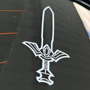 Op de afbeelding: Een witte vinyl sticker van een sierlijk zwaard, bevestigd op de zwarte achterruit van een voertuig. Het zwaardontwerp heeft een gestileerd lemmet, handvat en pareerstang, wat een decoratief embleem vormt.