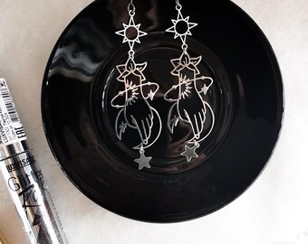 May include: A pair of silver dangle earrings with a celestial theme. Each earring features a sun, a crescent moon, a hand, and stars.