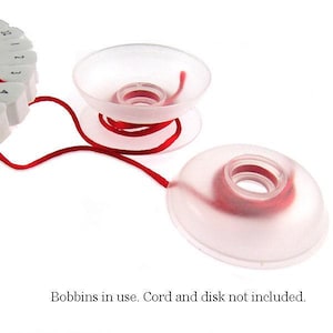 May include: Two clear plastic thread spool holders with red thread. The holders are round with a central opening. The text at the bottom of the image reads: "Bobbins in use. Cord and disk not included."