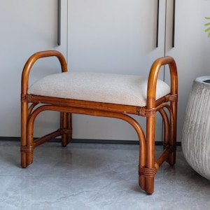 May include: A small, natural rattan bench with a light-colored upholstered seat. The bench has curved armrests and a woven design on the legs. The bench is approximately 30 inches long.