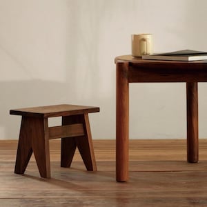 May include: A small, dark brown wooden stool with a rectangular seat and angled legs stands next to a round wooden table. The table features cylindrical legs and holds a light-coloured ceramic mug and a closed book on its surface.