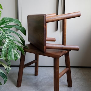 Puede incluir: Silla de madera artesanal, pequeña, con acabado marrón oscuro. Presenta un asiento cuadrado, cuatro patas robustas y un respaldo curvo. Diseño minimalista y moderno, ideal para la decoración del hogar.
