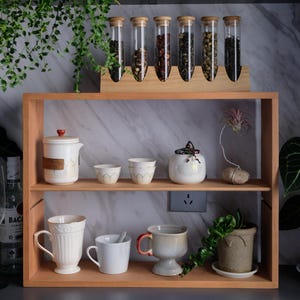 Puede incluir: Una estantería de madera exhibe artículos relacionados con el té. Seis tubos de vidrio con tapones de madera contienen varios tés. Debajo, una tetera, tazas y tazas están dispuestas. Una pequeña planta y una planta aérea decoran el conjunto.