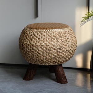 May include: A round, woven stool with a natural, light brown color. The stool has a woven top and a woven body, supported by three dark brown wooden legs. The stool is a decorative piece of furniture.