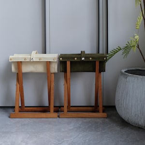 Könnte beinhalten: Zwei kleine Holzstühle mit Leinensitzen. Einer hat einen cremefarbenen Sitz, der andere einen olivgrünen Sitz. Beide haben Holzbeine und einen Griff. Die Hocker stehen vor einer grauen Wand. Ungefähre Höhe: 40 cm.