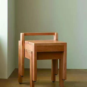 Peut inclure: Deux tabourets en bois, l'un empilé sur l'autre, avec une finition en bois naturel. Les tabourets ont un design simple et moderne avec des pieds carrés et une assise rectangulaire. Fond vert clair et sol en bois.