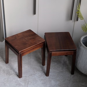 May include: Two small, square wooden stools in a dark brown hue. The stools feature a minimalist design with four straight legs and a flat top. They are positioned on a light grey tiled floor. The background is neutral.