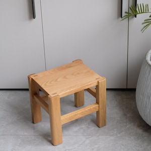 May include: A small, square wooden stool with a natural finish. The stool has four sturdy legs and a flat seat. The wood grain is visible, giving it a rustic and minimalist aesthetic. The stool is approximately 30 cm tall.