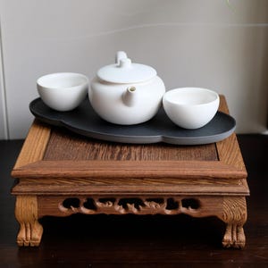 May include: A white ceramic teapot and two teacups are arranged on a dark gray tray, which is placed on a wooden stand with intricate carvings. The stand has four legs and is made of dark brown wood.