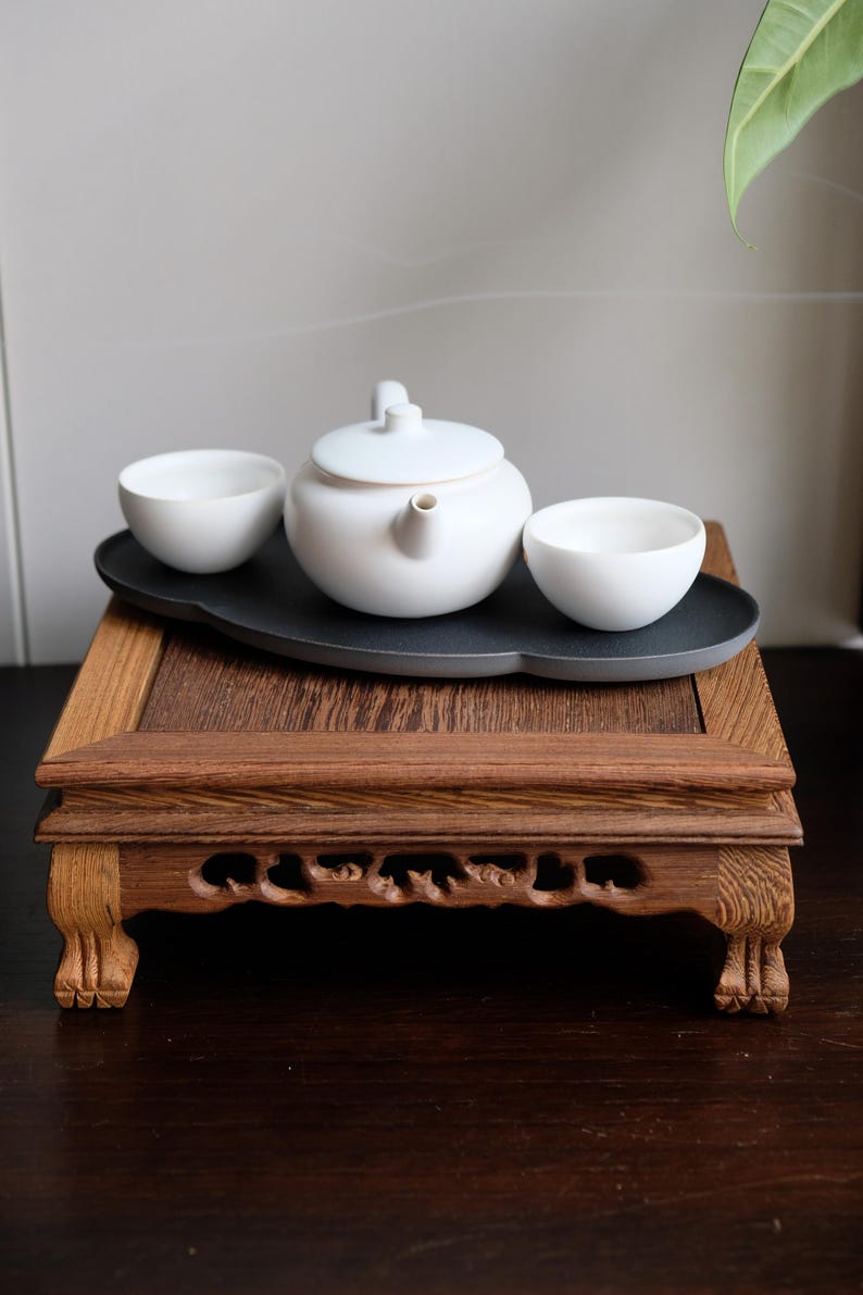 May include: A white ceramic teapot and two teacups are arranged on a dark gray tray, which is placed on a wooden stand with intricate carvings. The stand has four legs and is made of dark brown wood.