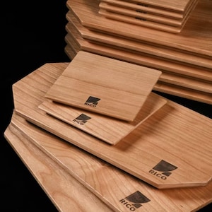 May include: A collection of wooden cutting boards and coasters. The boards are light brown with a natural wood grain and a geometric shape. Each board has the word "RICO" printed in black. The coasters are square and stacked.