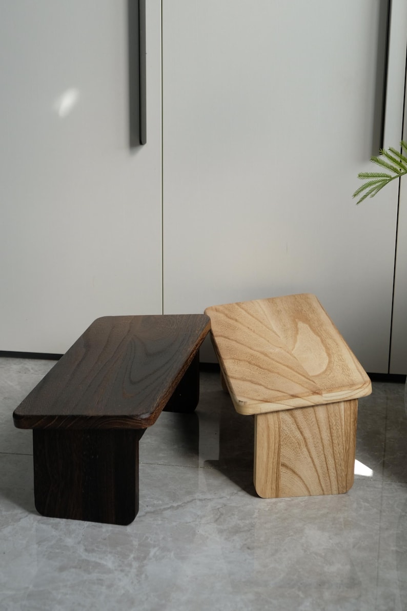 May include: Two small wooden stools. One is dark brown, the other is light brown. Both have a rectangular top and a square base. The stools are made of wood and have a natural finish. They are suitable for use as a footrest or small table.