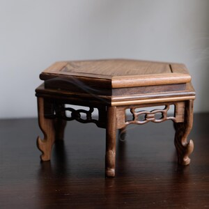 May include: A hexagonal wooden stand with intricate carvings and a dark brown finish. The stand has six legs and a flat top surface.