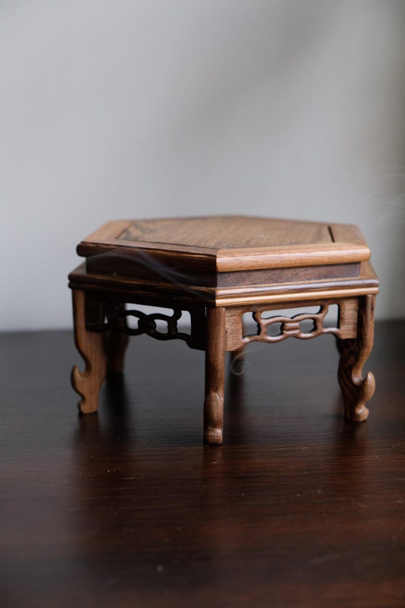 May include: A hexagonal wooden stand with intricate carvings and a dark brown finish. The stand has six legs and a flat top surface.