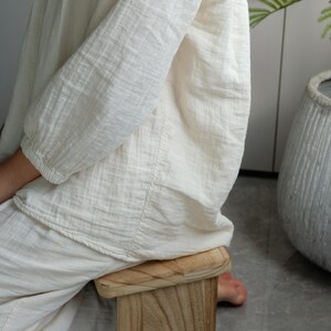 May include: A small, light-colored wooden stool with a rectangular seat and a square base. The stool is made of natural wood and has visible grain patterns. The person is wearing a cream-colored linen outfit.