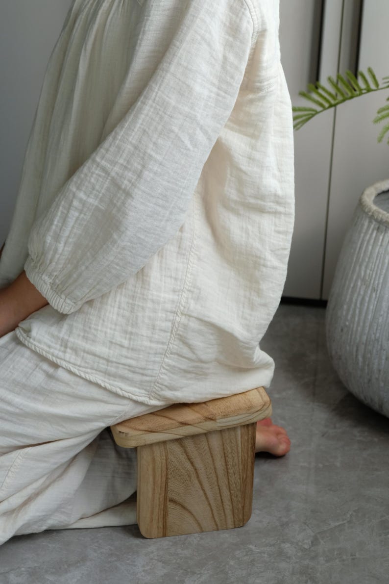 May include: A small, light-colored wooden stool with a rectangular seat and a square base. The stool is made of natural wood and has visible grain patterns. The person is wearing a cream-colored linen outfit.