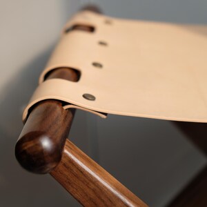 May include: A wooden folding stool with a light brown leather seat. The seat is secured with metal rivets.