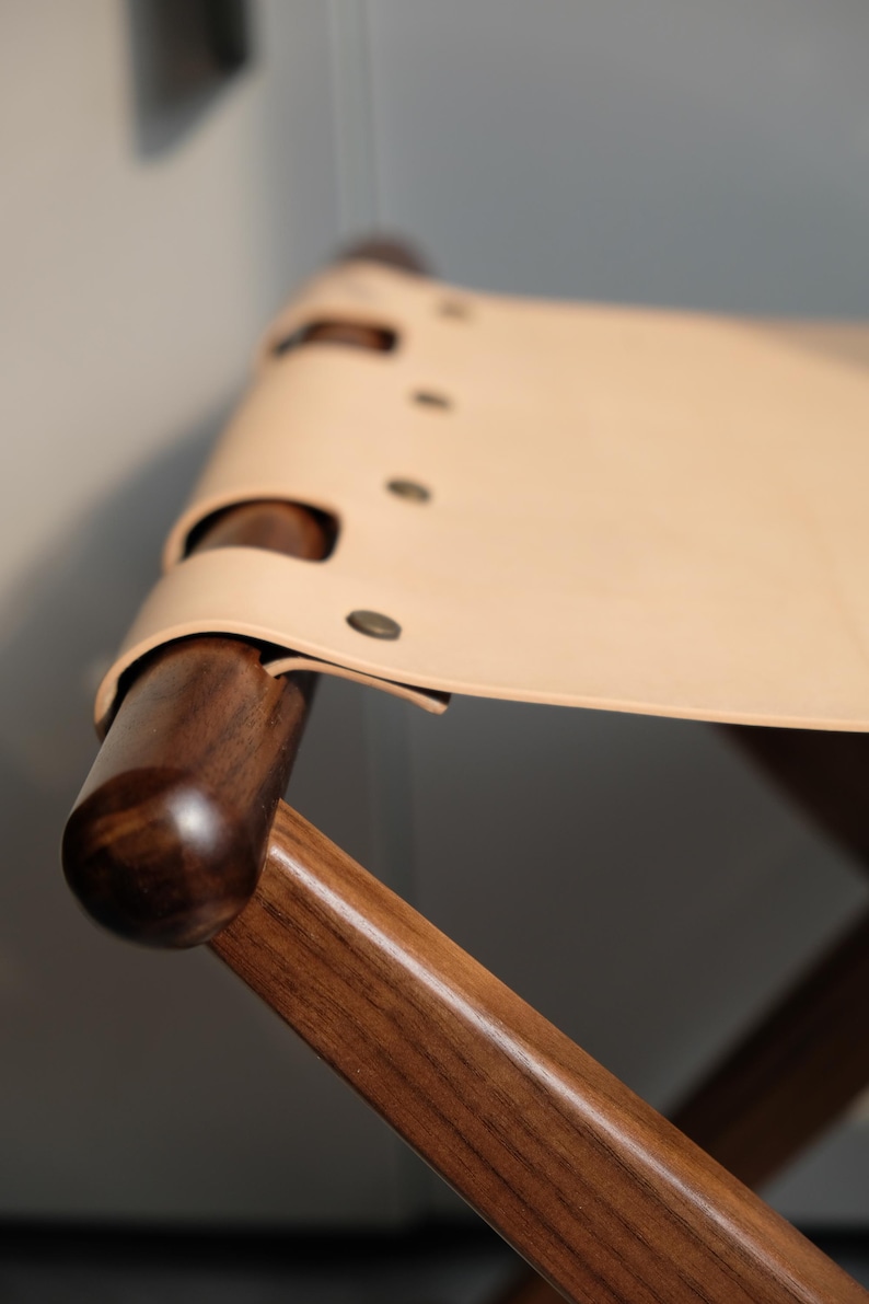 May include: A wooden folding stool with a light brown leather seat. The seat is secured with metal rivets.