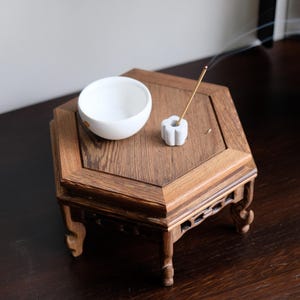 May include: A white ceramic bowl and a white ceramic incense holder with a burning incense stick on a hexagonal wooden table with carved legs.