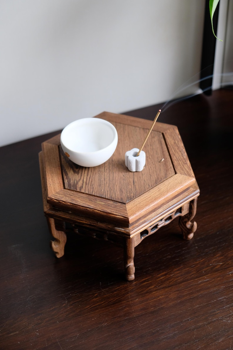 May include: A white ceramic bowl and a white ceramic incense holder with a burning incense stick on a hexagonal wooden table with carved legs.