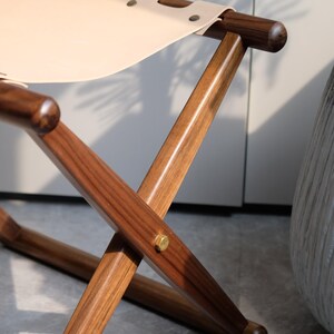 May include: A wooden folding chair with a light brown leather seat. The chair has a simple design with a cross-shaped frame and rounded legs.